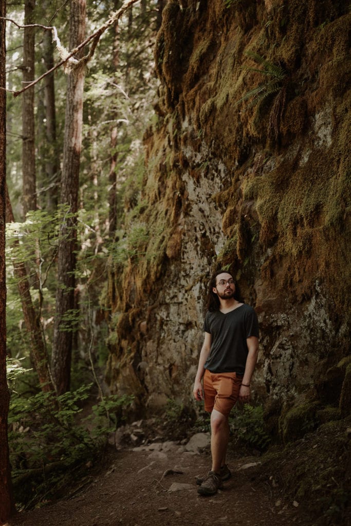 Hiker stands by large rock wall covered in moss. He has long hair and wears orange shorts