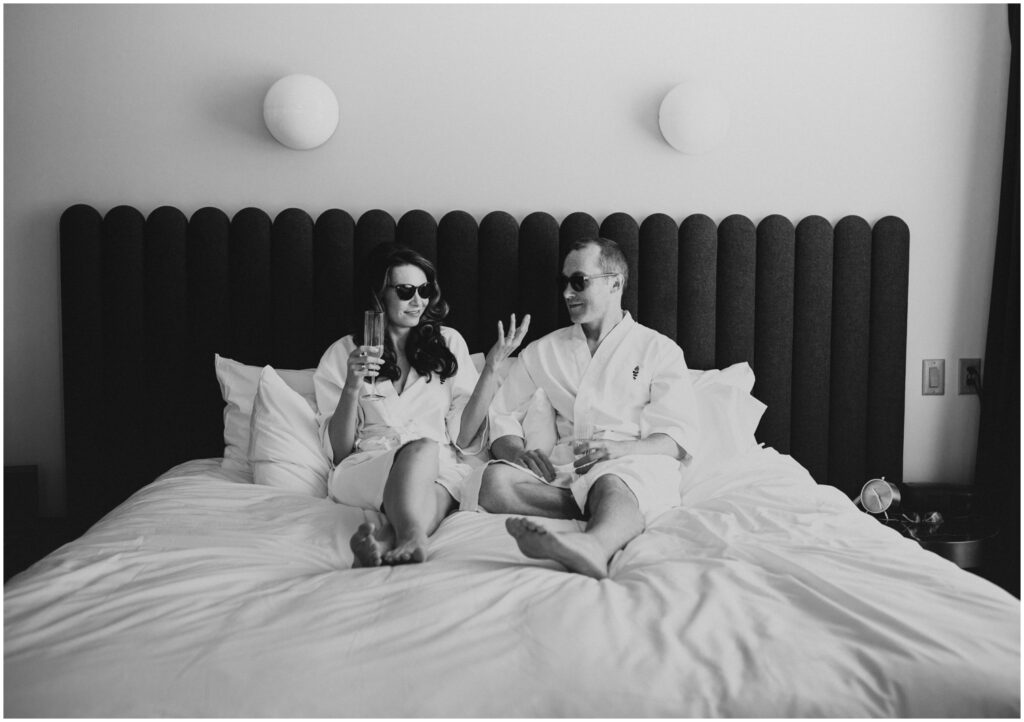 Bride and groom wear sunglasses and matching robes while lounging in bed before their wedding day
