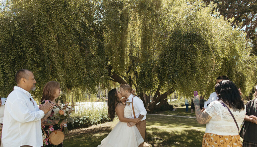 Bride and groom share a kiss at their ceremony under the shade with family and friends 