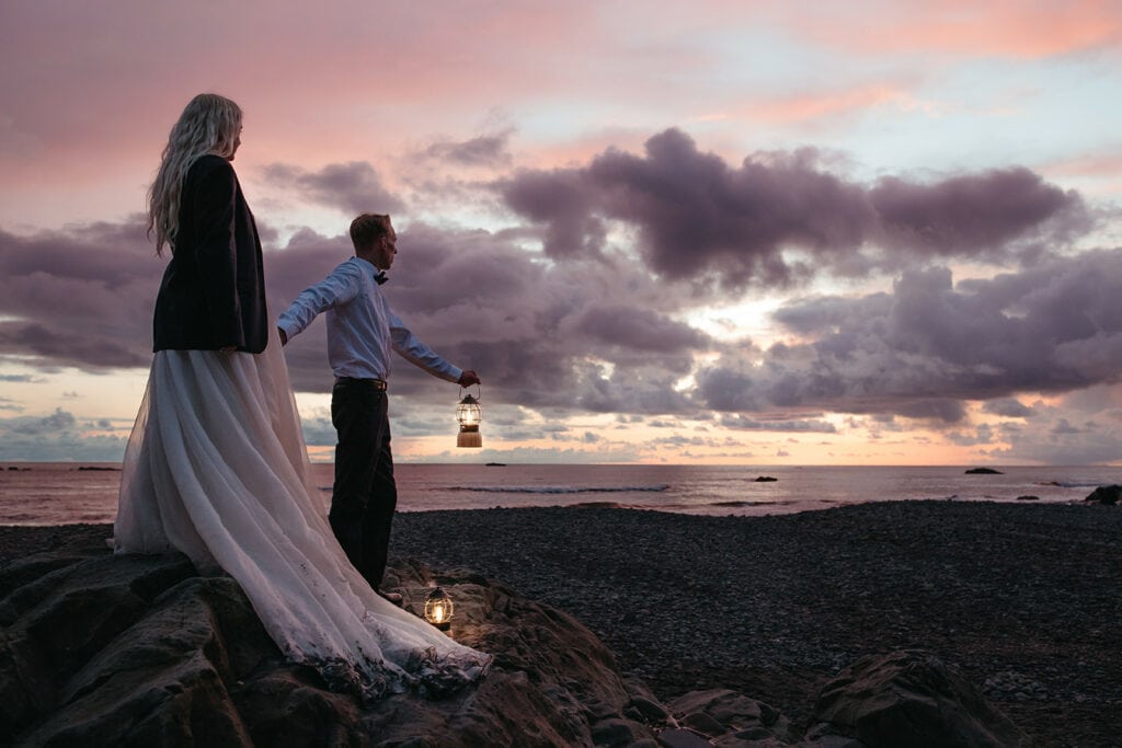 night time shot of bride and groom standing on the beach looking out into the water with the groom holding the brides hand and holding a lantern in the other hand 