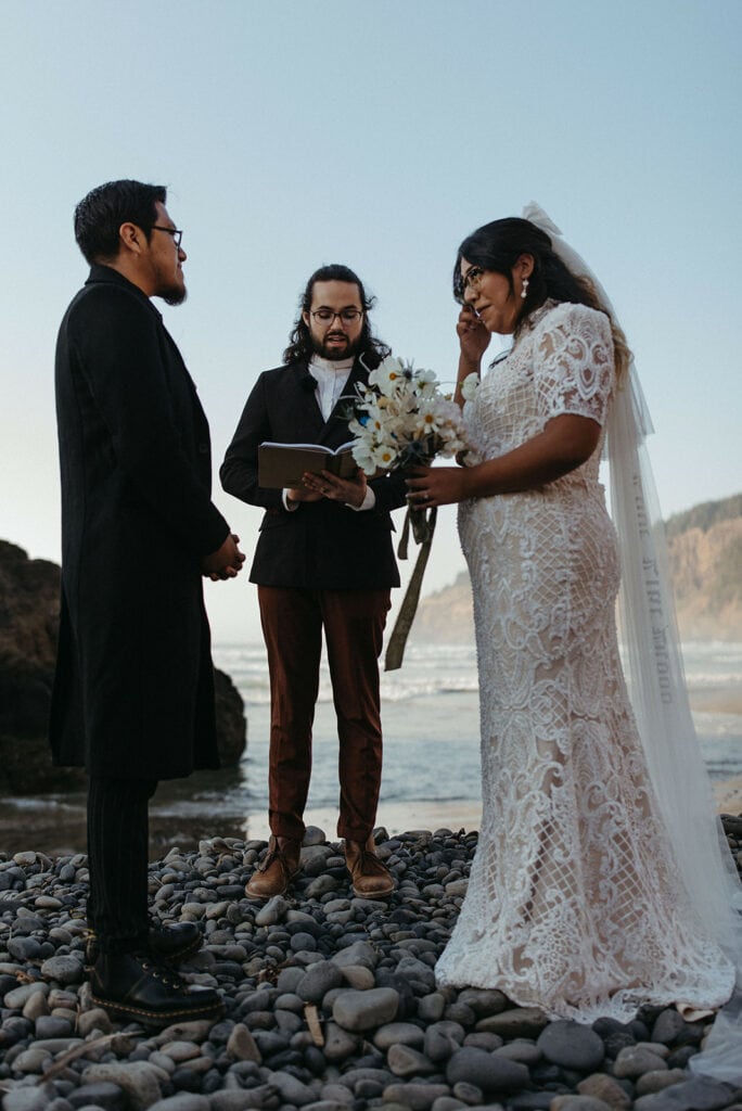bride and groom stand with officiant during beach wedding ceremony