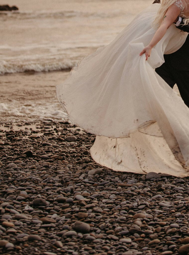 man holding woman on the beach while she lifts up her dirty wedding dress