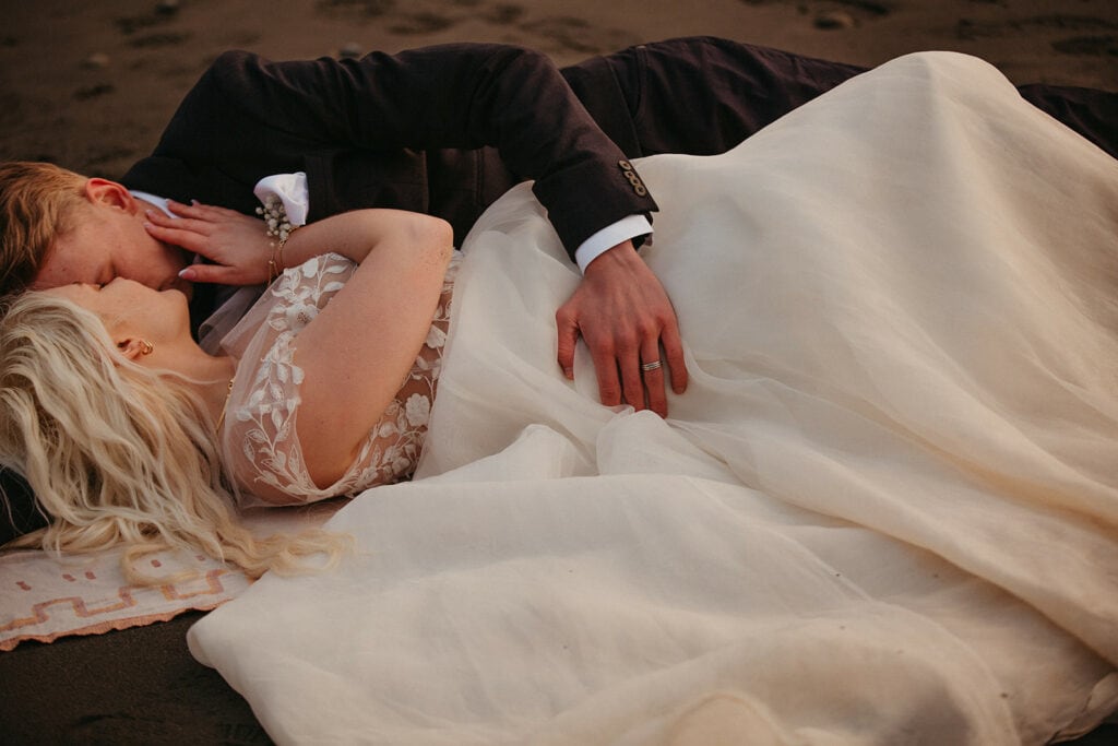 woman getting her wedding dress dirty as she lays on the beach with her husband
