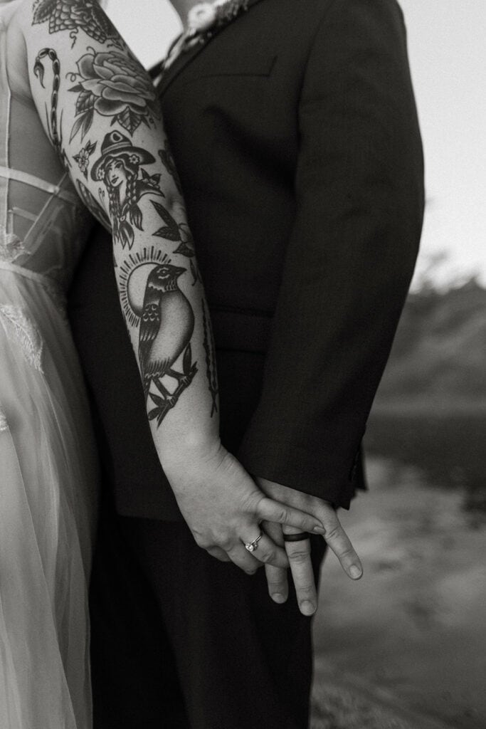 black and white photo with couple holding hands,  tattoos on wife's arm showcasing both rings 