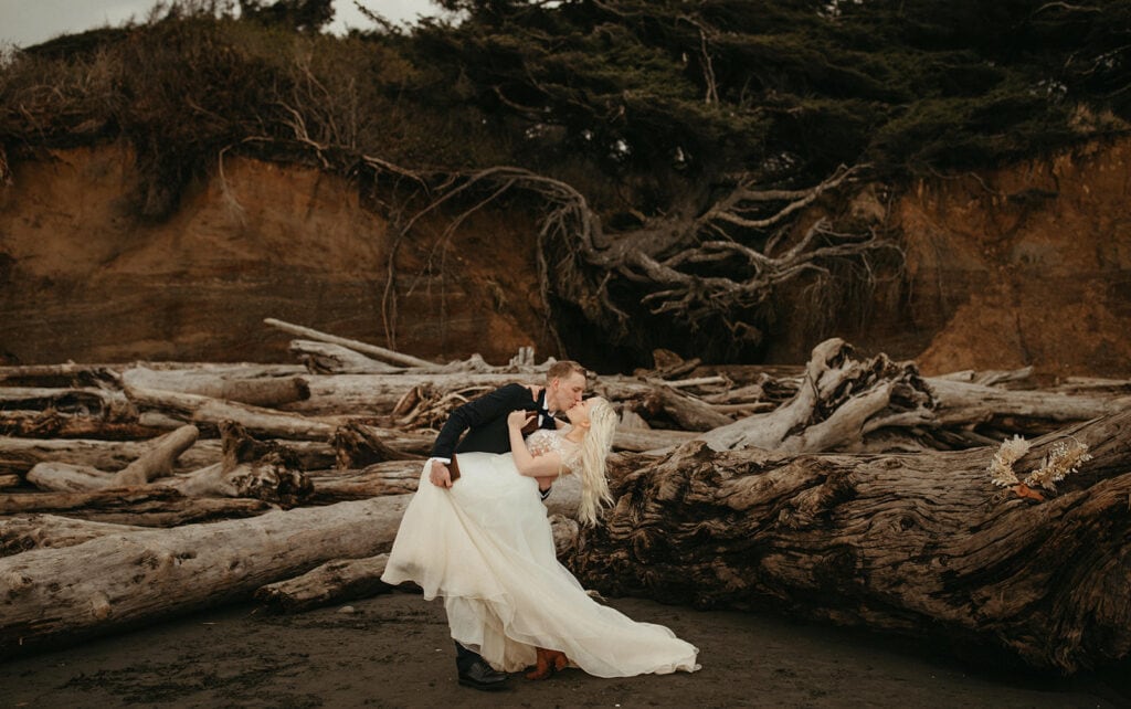 Groom bends his bride back for a kiss in front of Tree of Life at Kalaloch Beach
