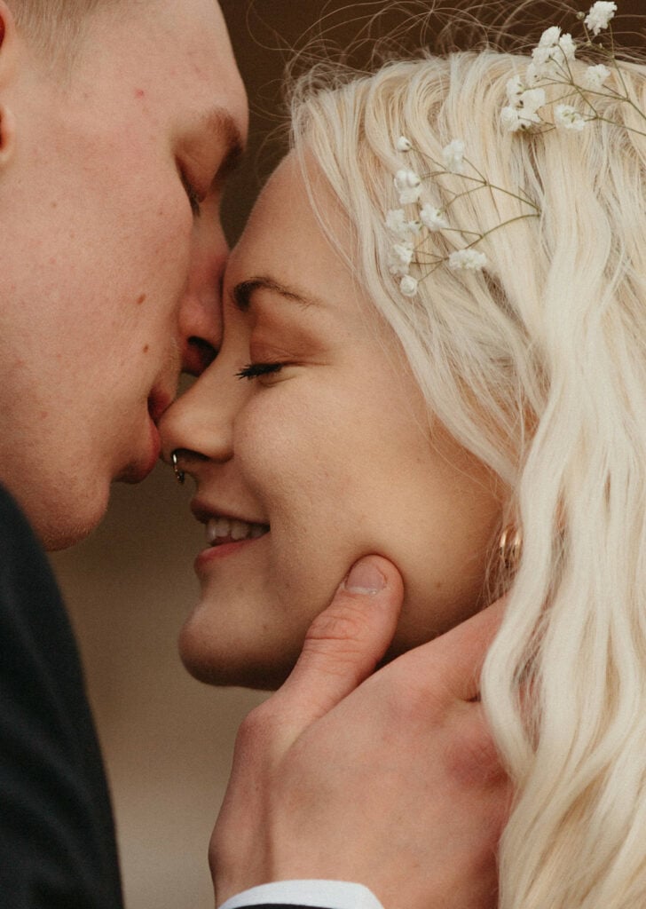 Bride smiles with flowers in her hair as groom gently kisses her nose