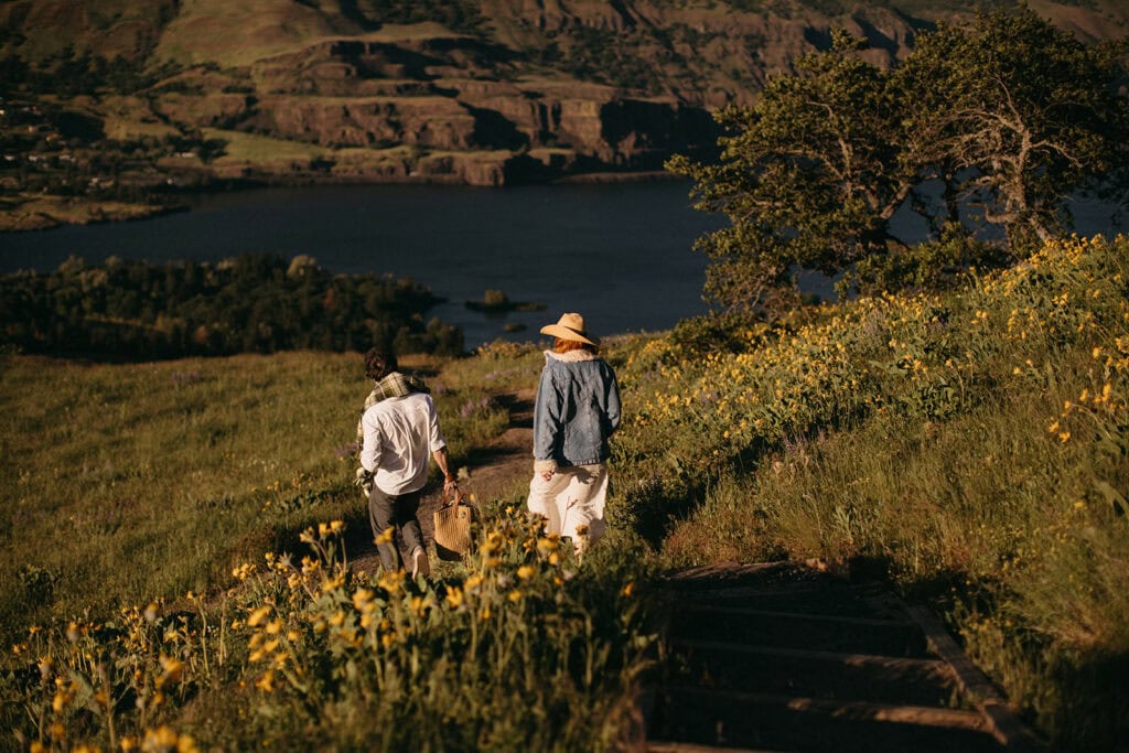 A couple walks down a dirt trail through a wildflower field overlooking the Columbia River for their adventure engagement photos